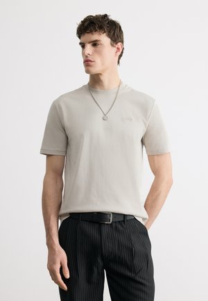 Jeune homme portant un t-shirt beige clair avec le logo « Boss », un pantalon noir à fines rayures, une ceinture noire et un collier pendentif argenté, debout devant un fond blanc.