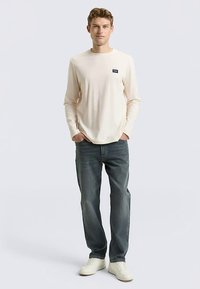 Beiges Langarmshirt mit kleinem Logo, kombiniert mit blauen Jeans und weißen Sneakern. Model steht vor hellem Hintergrund.