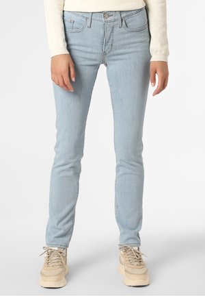 Personne portant un jean skinny bleu clair, des baskets beiges et un haut à manches longues crème, debout devant un fond clair.