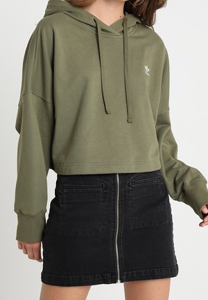 Kobieta nosi oliwkową krótką bluzę z kapturem ze sznurkami oraz czarną mini spódnicę jeansową z zamkiem błyskawicznym z przodu i kieszeniami.