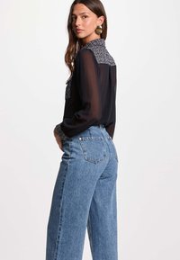 Blusa negra transparente con cuello y puños texturizados, combinada con jeans de cintura alta color azul claro. La modelo está posando de lado, mostrando los detalles del atuendo.