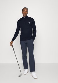 Navy zip-up sweater with logo, coupled with dark pants and white golf shoes. Model holding a golf club, standing on a neutral background.
