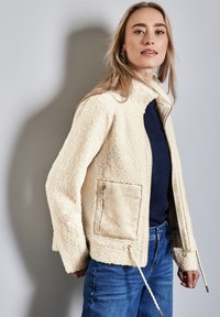 Veste en polaire crème avec un col haut, une poche zippée et des cordons à la taille. Associée à un denim bleu, mettant en avant un style décontracté.