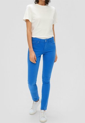 Person iført hvid kortærmet skjorte, lyseblå slim-fit jeans og hvide sneakers, stående mod en ensfarvet baggrund.