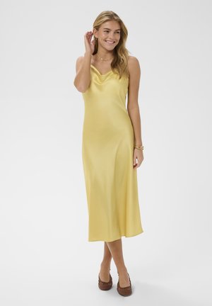 Mosolygó nő sárga szatén ujjatlan midi ruhában, barna szövött magas sarkú cipőben, arany nyaklánccal, karkötővel és fülbevalóval fehér háttér előtt.