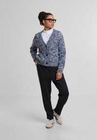 Gebreide blauw-witte cardigan met een V-hals en vijf knopen, gecombineerd met zwarte broek en een witte blouse, gedragen met casual sneakers.