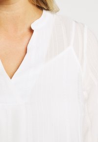 Blusa bianca con un delicato motivo a righe verticali, scollatura a V e una texture leggera. Il tessuto appare morbido e leggermente trasparente.