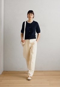 Navy knitted cardigan with short sleeves, cream trim, beige high-waisted pants, and light footwear. Simple design against a light background.