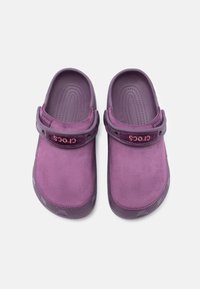 Une paire de sabots Crocs violets avec un dessus en imitation suède et une sangle arrière réglable, vue d'en haut sur un fond blanc.