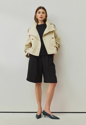 Frau trägt beige kurz geschnittene Trenchjacke, schwarzes Hemd, lockere schwarze knielange Shorts und dunkle spitze flache Schuhe steht an einer Wand.