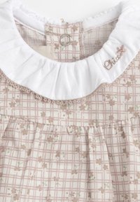 Robe rose clair avec un col blanc à volants. Elle présente un motif quadrillé avec de petites impressions florales, la texture du tissu est en coton. La fermeture à l'arrière se fait par un bouton.