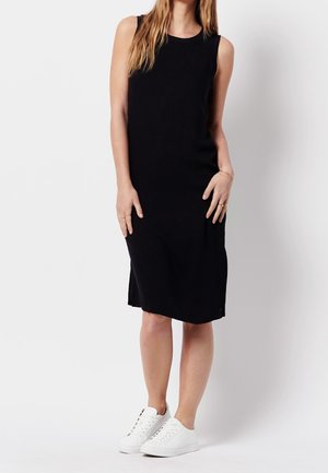 Femme debout portant une robe noire sans manches mi-longue et des baskets blanches sur un fond blanc uni.