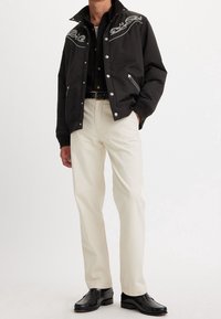 Svart bomberjacka med silverbroderade detaljer, fickor med dragkedja, i kombination med krämfärgade byxor och svarta läderskor.