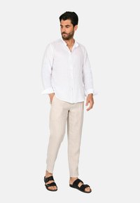 Camicia bianca in lino con bottoni, vestibilità rilassata. Pantaloni in lino beige chiaro e sandali neri. Colori neutri e trame morbide, adatti per il clima caldo.