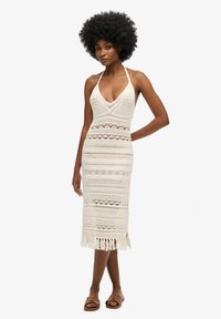 Strickcrochet-Kleid in Off-White mit Halterneck, figurbetontem Schnitt, komplizierten Mustern und Quastenabschlüssen. Flache Sandalen getragen.