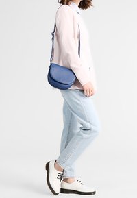 Blå lädercrossbody-väska med en halvcirkelform, slät yta och justerbar rem. Bärs med ljus denim och vita skor.