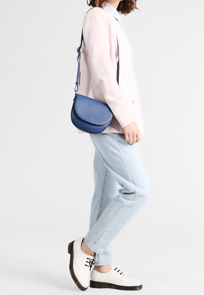Modrá kožená crossbody taška v polokruhovém tvaru, hladkým povrchem a nastavitelným popruhem. Nosená s lehkými džíny a bílými botami.
