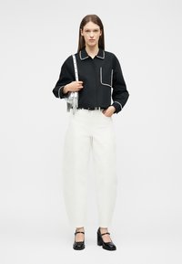 Mujer con una camisa negra de manga larga con ribete blanco, pantalones blancos de talle alto, tacones negros y un bolso plateado colgado del hombro.