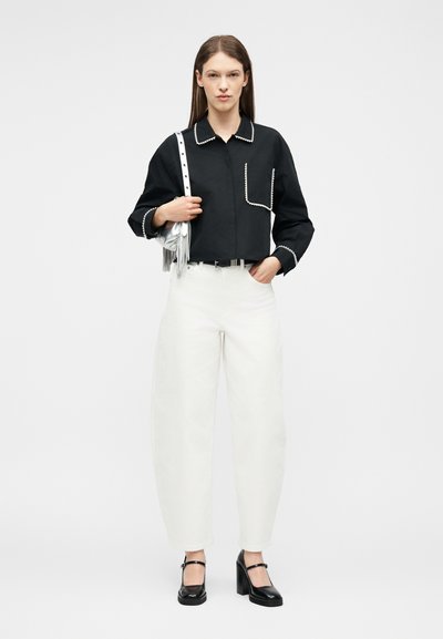 Γυναίκα με μαύρο πουκάμισο μακρυμάνικο με άσπρο περίγραμμα, ψηλόμεσο άσπρο παντελόνι, μαύρα τακούνια και κρατώντας μια ασημί τσάντα στον ώμο της.