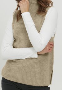 Beige-ribbineulottu poololiivimalli, jossa on sivuhalkiot, puettuna valkoisen pitkähihainen paidan päälle. Liivissä on teksturoitu pinta ja vartalonmyötäinen muotoilu.