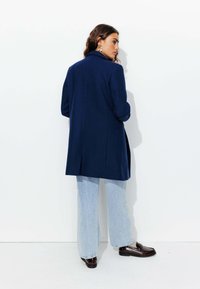 Manteau en laine bleu marine au design ajusté, avec des revers crantés et une silhouette élégante, associé à un jean large bleu clair et des mocassins foncés.