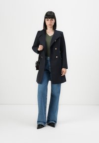 Manteau en mélange de laine bleu marine avec des boutons dorés à double boutonnage, un large col et des poches latérales, porté sur un haut vert et un pantalon en jean bleu à jambes larges.