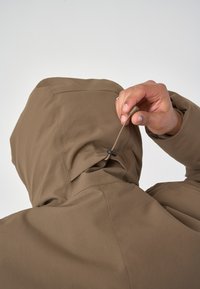 Braune Kapuzenjacke mit glatter Textur. Eine Hand stellt den Knebel an der Kapuze ein und zeigt damit die verstellbaren Designelemente und die sauberen Nähte.