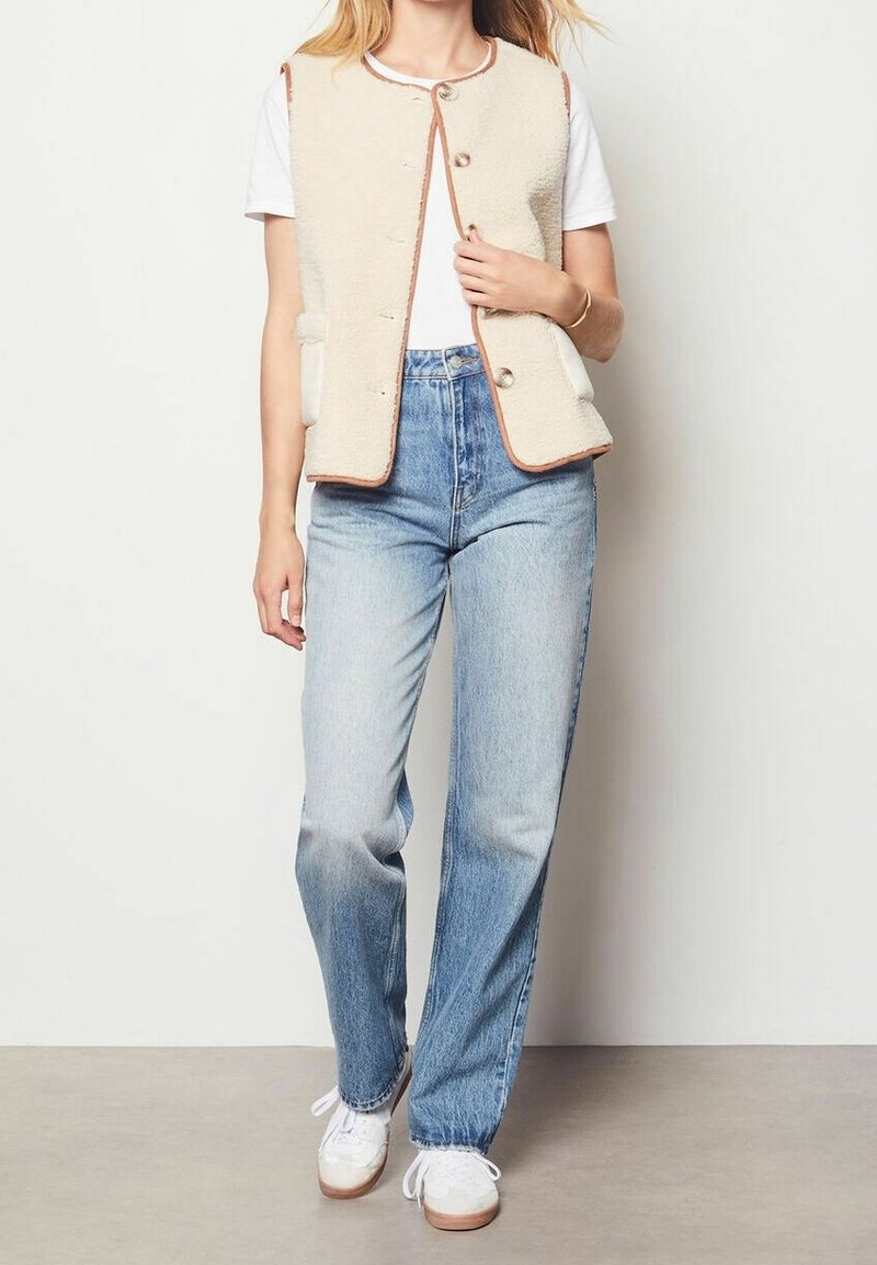 Personne portant un t-shirt blanc, une veste en polaire crème boutonnée avec une bordure marron, un jean taille haute bleu clair et des baskets blanches, debout.