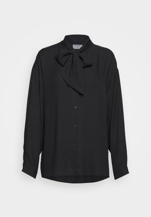 Nainen pukeutunut mustaan pitkähihainen puseroon, jossa on rusetti, korkeavyötäröiset, leveälahkeiset mustat housut, mustat sandaalit ja pitää sisätiloissa mustaa clutch-laukkua.