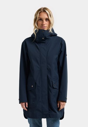 Femme portant un long imperméable bleu marine avec un col montant et des poches avant à boutons-pression, debout devant un fond clair uni.