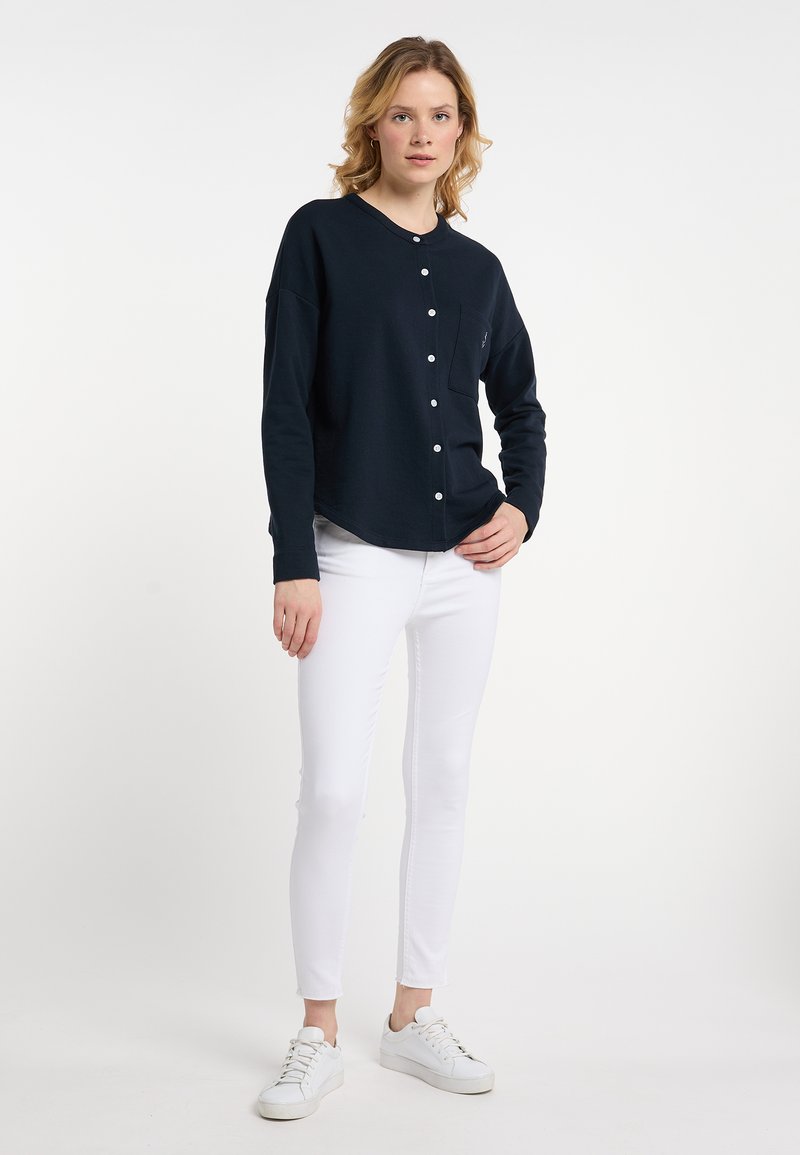 Camicia blu navy a maniche lunghe con bottoni e tasca, orlo arrotondato, realizzata in tessuto morbido, abbinata a pantaloni bianchi aderenti e sneakers bianche.