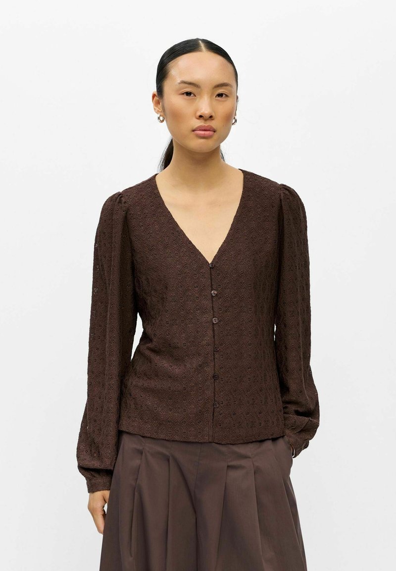Femme aux cheveux foncés attachés porte un chemisier marron foncé à manches longues boutonné en tissu texturé et une jupe plissée marron.