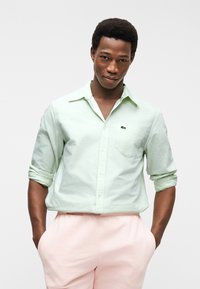 Homme les mains dans les poches, portant une chemise boutonnée vert clair à manches retroussées et un pantalon rose pâle, sur un fond blanc uni.