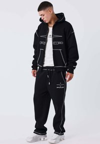 Joven mirando hacia abajo, vistiendo una sudadera negra con cremallera y pantalones de chándal negros a juego con costuras blancas y zapatillas blancas sobre un fondo liso.