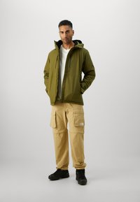 Jachetă impermeabilă de culoare verde măsliniu cu glugă, pantaloni cargo bej cu buzunare mari și bocanci negri de drumeție. Design simplu cu detalii în contrast.