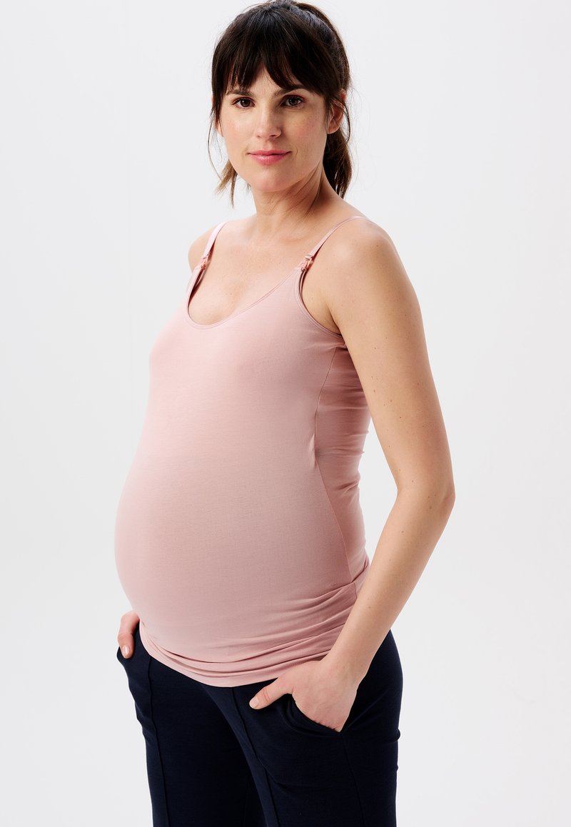 Zwangere vrouw met donker haar in een roze tanktop en marineblauwe broek staat met haar handen in haar zakken tegen een witte achtergrond.