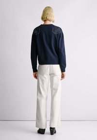 Felpa navy con design decorativo di strass sulle spalle, abbinata a pantaloni bianchi a gamba larga e scarpe con tacco verdi; sfondo semplice.