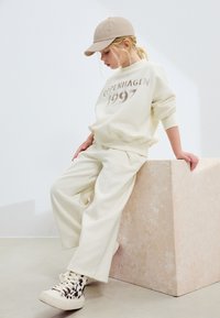 Tenue couleur crème comprenant un sweat-shirt ample avec l'inscription "COPENHAGEN 1997" en imprimé métallique et un pantalon assorti. Casquette et baskets à motifs incluses.