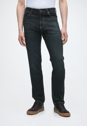 Homme portant un jean slim foncé et des baskets noires avec des semelles marron, debout devant un fond neutre.