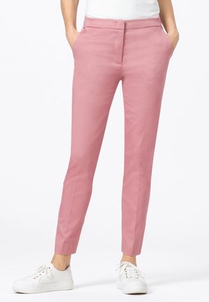 Femme portant un pantalon rose slim avec des poches latérales et des baskets blanches, se tenant devant un fond blanc uni.