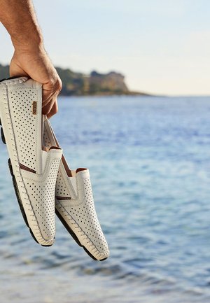 Hvide perforerede slip-on sko med brune detaljer, med en syet kant og sorte gummisåler, taget imod en havudsigt.