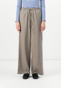 Persoon in losse beige wijde broek met een trekkoord in de taille, gestreepte blauw-witte lange mouwen top, en zwarte schoenen, staande op een witte vloer.
