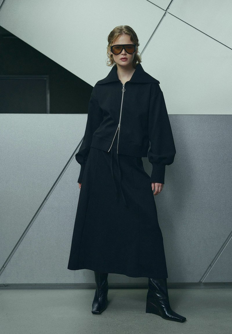 Femme debout portant une veste noire zippée oversize, une longue jupe noire, des bottes compensées noires et de grandes lunettes de soleil marron devant un mur géométrique.