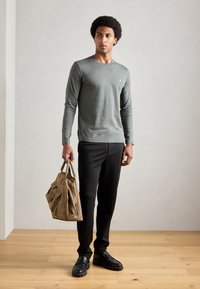Camicia a maniche lunghe verde oliva, pantaloni neri e mocassini neri. Borsa marrone con diverse tasche. Design minimalista, texture liscia.