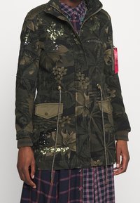 Maskovací parka s květinovým vzorem, zdobená flitry, se stahovací šňůrkou v pase a dvěma předními kapsami. Látka má strukturovaný povrch.