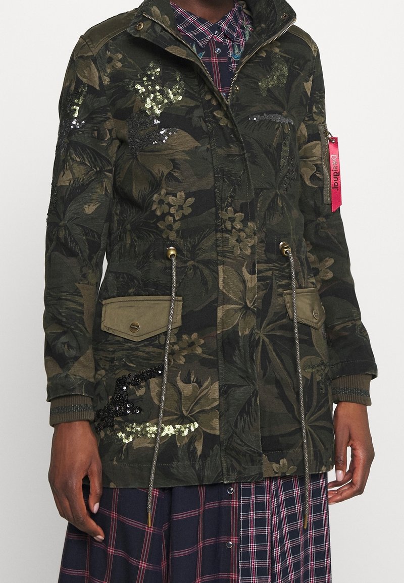 Maskovací parka s květinovým vzorem, zdobená flitry, se stahovací šňůrkou v pase a dvěma předními kapsami. Látka má strukturovaný povrch.