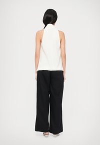 Muguras skats uz balta topu ar kakla siksniņu, kas kombinēts ar vaļējām melnām biksēm. Audums šķiet gluds, bez redzamām rakstām vai akcēm.