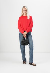 Sudadera roja, jeans azules y bolso negro texturizado. Zapatos con estampado de leopardo. Modelo de pie contra un fondo blanco liso.