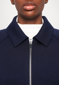Námornícka modrá bunda na zips s golierom, obsahujúca strieborný zips a ribovanú textúru, priliehavý strih a čistý, minimalistický dizajn.