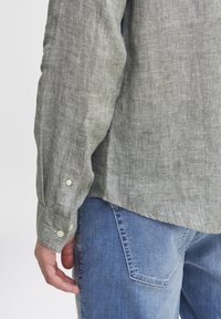 Camicia di lino verde chiaro con maniche lunghe, polsini a bottoni e una vestibilità rilassata, indossata sopra dei jeans blu in denim.